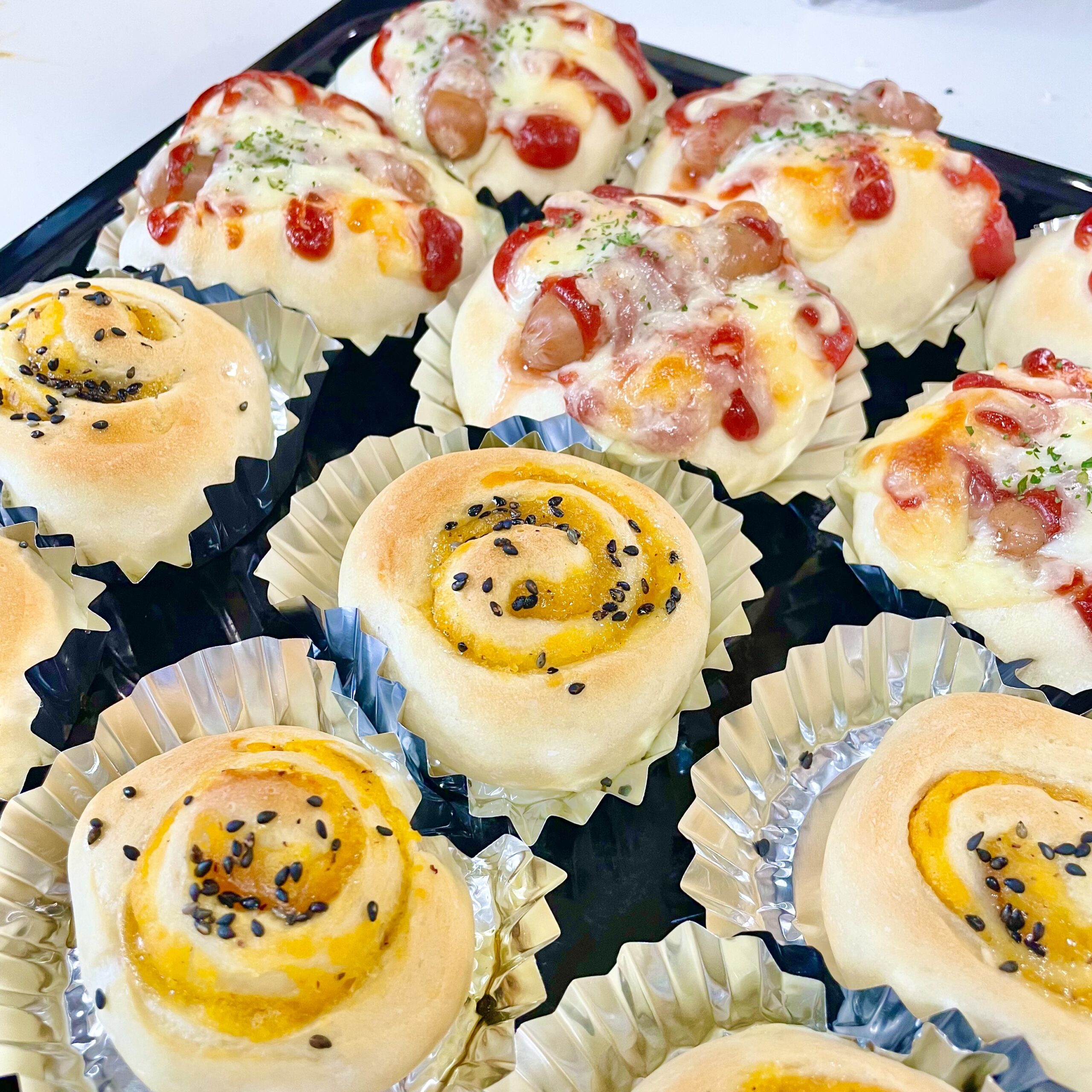 焼きたてのウインナーパンとさつまいもロールパンが並んだトレイの写真。ふわふわ食感の生地にチーズやごまがトッピングされ、彩りよく仕上がっている。