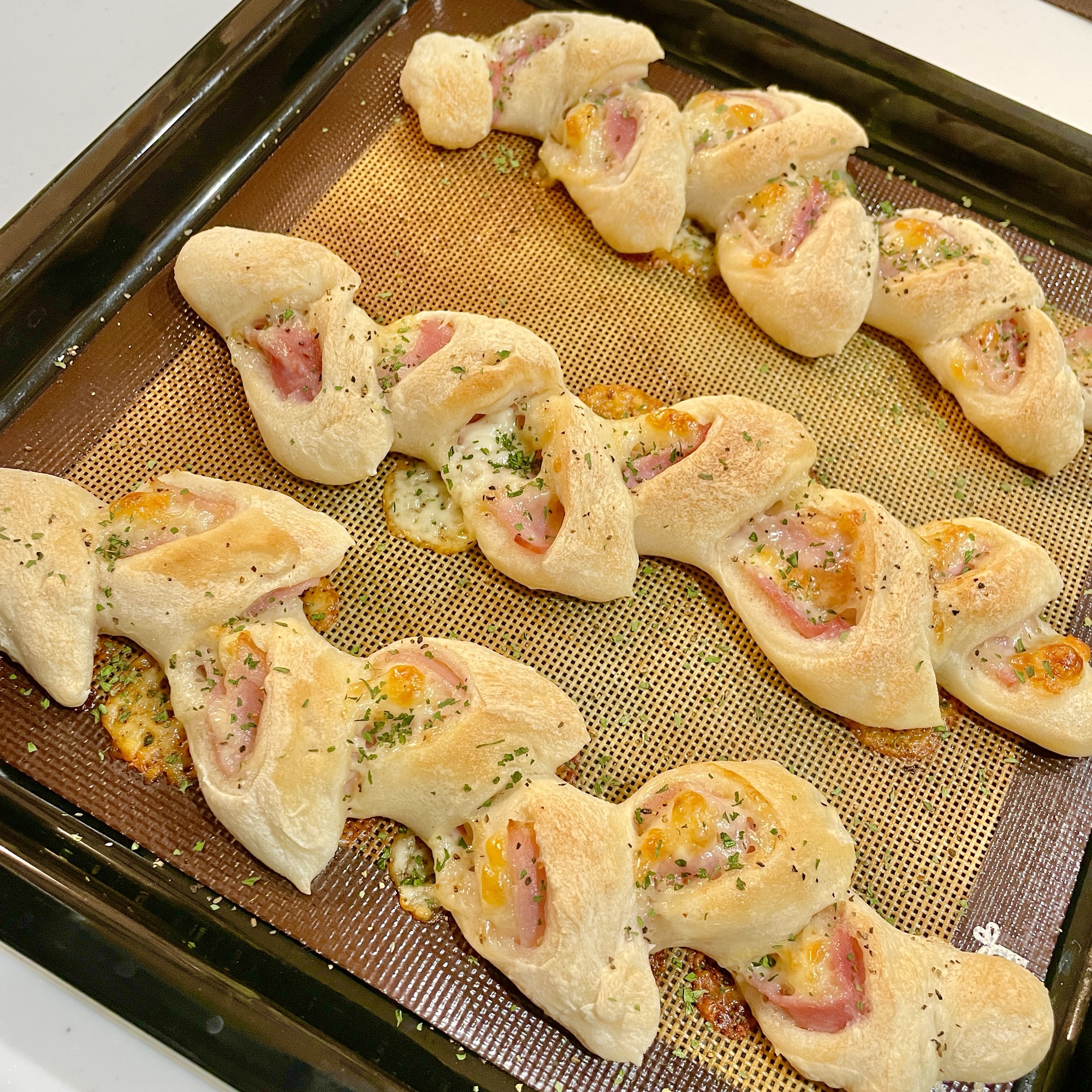 焼き立てのベーコンエピがオーブンシートの上に並んでいる。黄金色に焼けた生地からベーコンとチーズが覗き、表面にはパセリがトッピングされている。カリッとした焼き目とふんわりしたパン生地が美味しそうな手作りベーコンエピの写真。